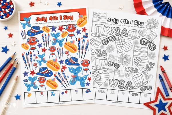 Two July 4th I Spy printable worksheets on a white tabletop, one colorful and one black-and-white coloring page, surrounded by patriotic red, white, and blue craft supplies, crayons, pencils, and star decorations.